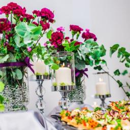 Niku Bowling Monika Gościniak - Dekoracja stołu z kwiatami w wazonach z fioletowymi wstążkami, świecami w szklanych lampionach na srebrnych świecznikach i przekąskami na srebrnej tacy, widoczne z bliska.