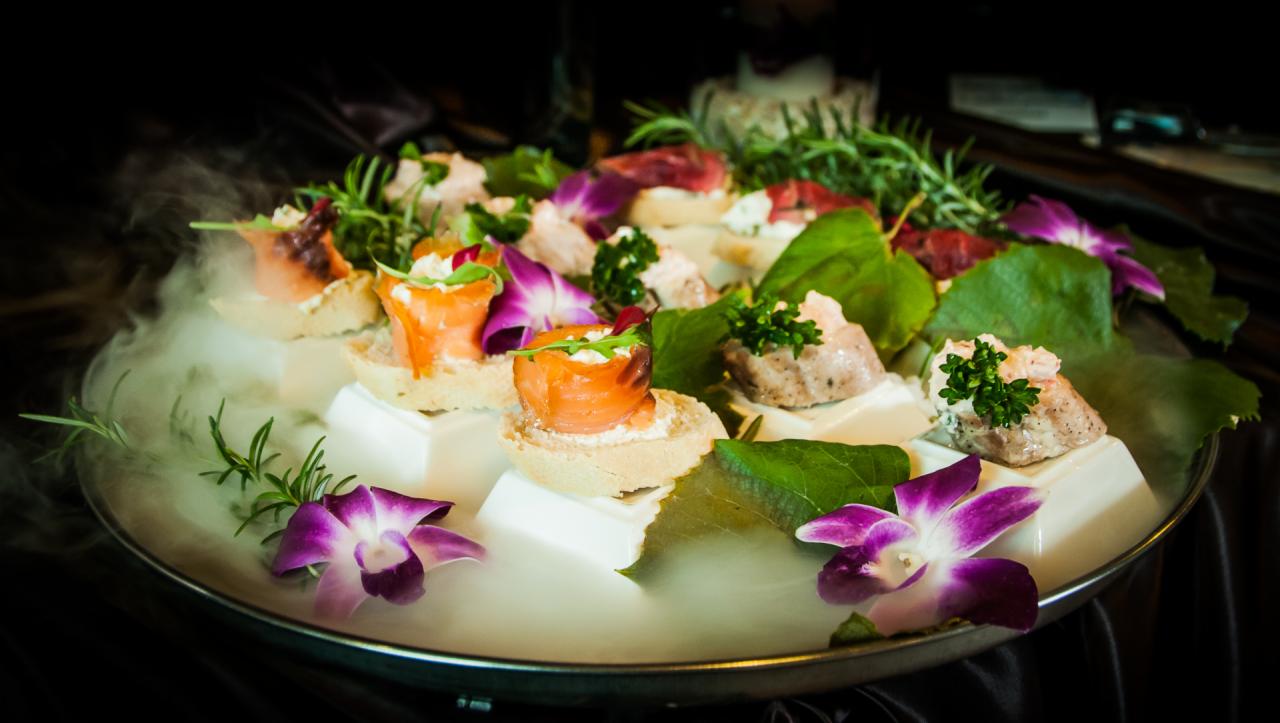 Ekskluzywne finger food na srebrnej tacy, z dekoracją z kwiatów orchidei, z efektem suchego lodu, na białych kostkach.