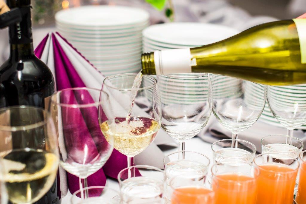Nalewanie białego wina z butelki do kieliszka na stole z ułożonymi talerzami, serwetkami i sokiem pomarańczowym w małych szklankach.