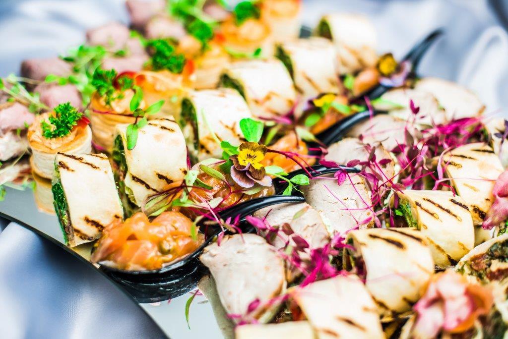 Elegancko zaaranżowana taca z różnorodnymi przekąskami cateringowymi, w tym roladki z grilla, mini tartaletki i kawałki wędzonego łososia, udekorowane świeżymi ziołami i jadalnymi kwiatami...