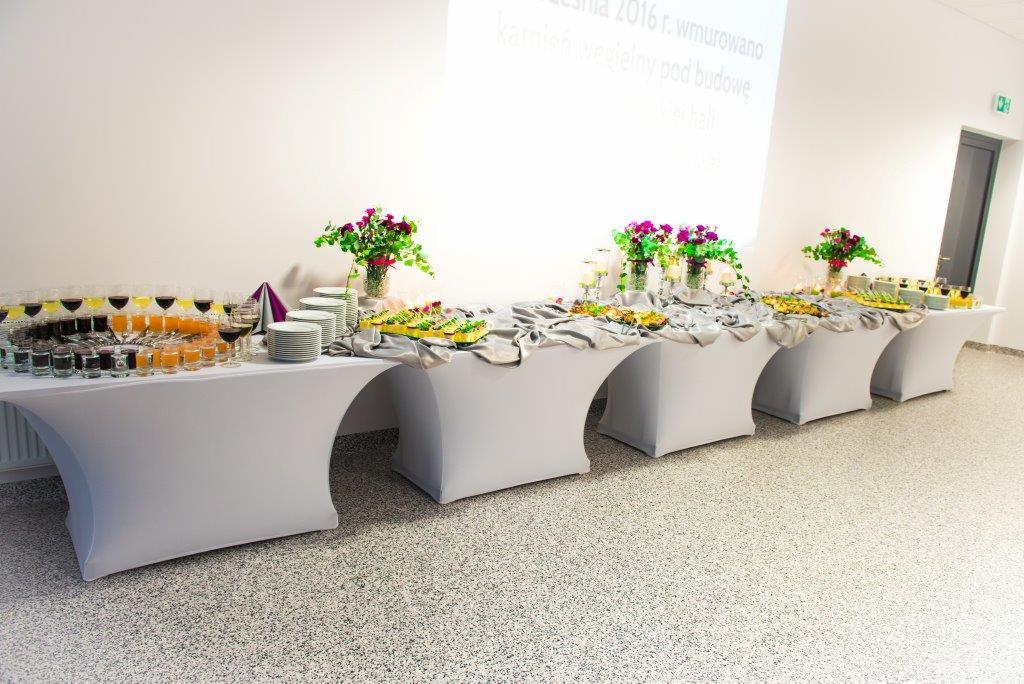Stół cateringowy z białym obrusem, na którym znajdują się napoje w szklankach, talerze, przekąski, kwiaty w wazonach i srebrna dekoracyjna tkanina. W tle biała ściana z napisem i drzwi.
