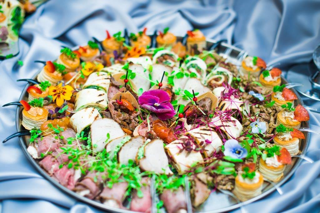 Dekoracyjna srebrna taca z różnorodnymi przekąskami: roladki z grillowanej cukinii, mini tortille z mięsem, roladki z wędlin, mini tartaletki z kremem, udekorowane świeżymi kwiatami i mikro-ziołami...