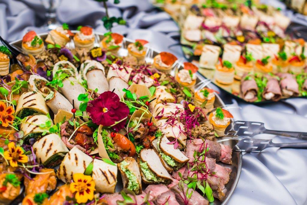 Dekoracyjnie podane przekąski na srebrnej tacy, w tym roladki z grilla, mini tortille z mięsem i warzywami, oraz kruche babeczki z pastą i pomidorem, ozdobione kwiatami i mikro-ziołami na tle...