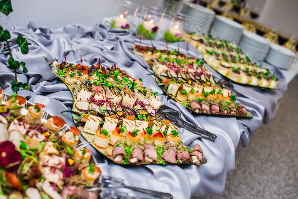 Stół cateringowy z różnorodnymi przekąskami na srebrnej, marszczonej tkaninie, widoczne mini tortille, roladki z mięsa, kanapeczki, dekoracje z ziół i kwiatów, w tle stosy białych talerzy i świeczki.