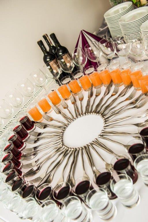 Dekoracyjne ułożenie kieliszków z czerwonym, pomarańczowym i bezbarwnym płynem, butelek wina i sztućców na srebrnej tacy, tworzące efektowną kompozycję.