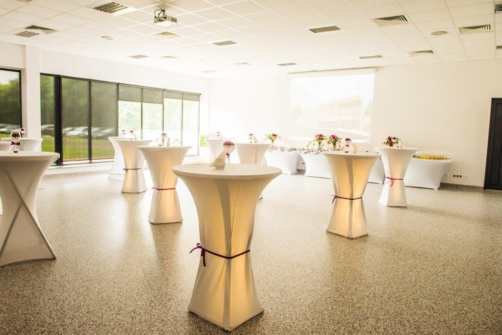 Sala bankietowa z wysokimi, podświetlanymi stolikami koktajlowymi, dekorowanymi bordowymi wstążkami, ustawionymi na parkiecie w jasnym wnętrzu z oknami i projektorem.