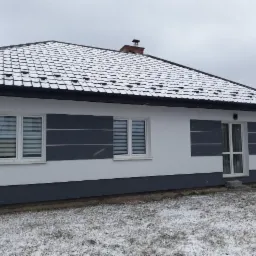 Nowoczesny dom jednorodzinny z białą elewacją, szarymi akcentami i śniegiem na dachu w zimowej scenerii.