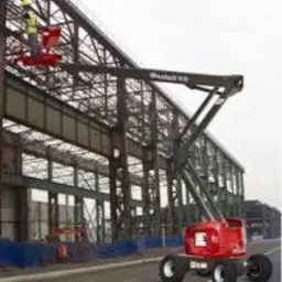 Czerwona platforma robocza Manlift 85 z wysięgnikiem teleskopowym unosi dwóch pracowników w kamizelkach odblaskowych na tle konstrukcji stalowej hali.