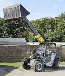 Kompaktowa ładowarka teleskopowa z podniesioną łyżką, widoczna na tle muru i zieleni, w słoneczny dzień.