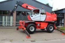 Czerwona koparko-ładowarka teleskopowa marki Manitou z wysuniętymi podporami, zaparkowana na kostce brukowej przed budynkiem z szarym dachem i ciemnymi drzwiami garażowymi.