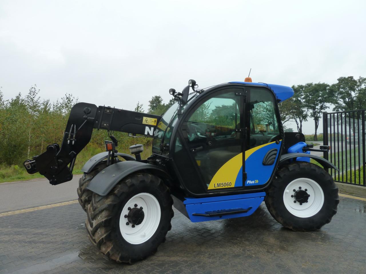 Niebiesko-czarna koparko-ładowarka teleskopowa New Holland LM5060 Plus z wysuniętym ramieniem, widok z boku na szarym bruku.