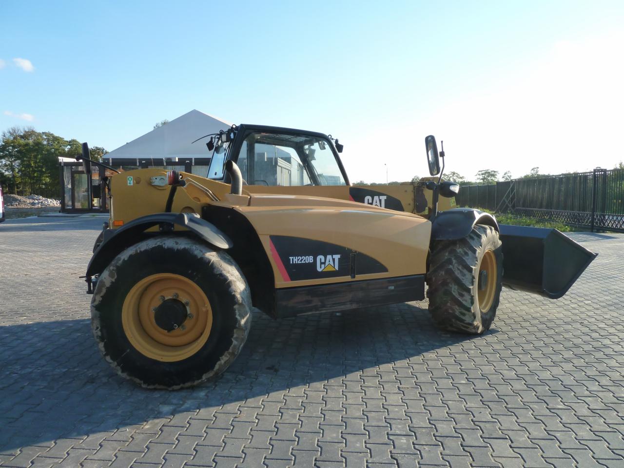 Żółta koparko-ładowarka CAT TH220B zaparkowana na kostce brukowej w Jawczycach, widok z boku, z łyżką skierowaną w prawo.