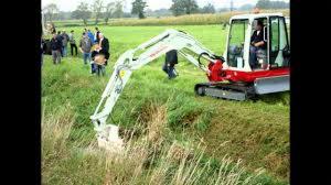 Mała, czerwono-biała koparka gąsienicowa na nierównym terenie trawiastym, z grupą osób obserwujących pracę w tle. Operator w kabinie, ramię koparki opuszczone nad wykopem.