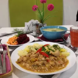 Zestaw obiadowy: gulasz z ziemniakami posypany koperkiem i papryczką chili, zupa w niebieskiej misce, surówka z buraków i marchewki, kompot w szklance, sztućce w koszyczku na stole w kratę.