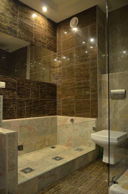 Wykończona łazienka z brązowymi i beżowymi płytkami imitującymi marmur, widoczny brodzik z odpływami kwadratowymi w podłodze, toaleta oraz szklana kabina prysznicowa.