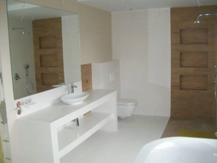 Nowoczesna łazienka z białymi płytkami, umywalką nablatową i drewnianymi elementami dekoracyjnymi w strefie prysznica, widoczne niedokończone prace wykończeniowe przy ścianie.