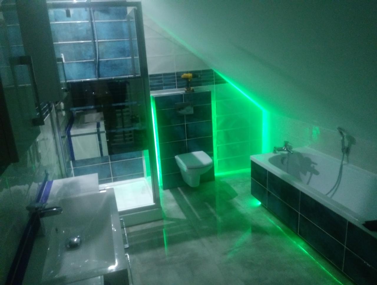 Wykończona łazienka na poddaszu z kabiną prysznicową, umywalką, toaletą i wanną, podświetlona zielonymi ledami wzdłuż krawędzi ścian i wanny. Na ścianie nad toaletą widoczna wiertarka.