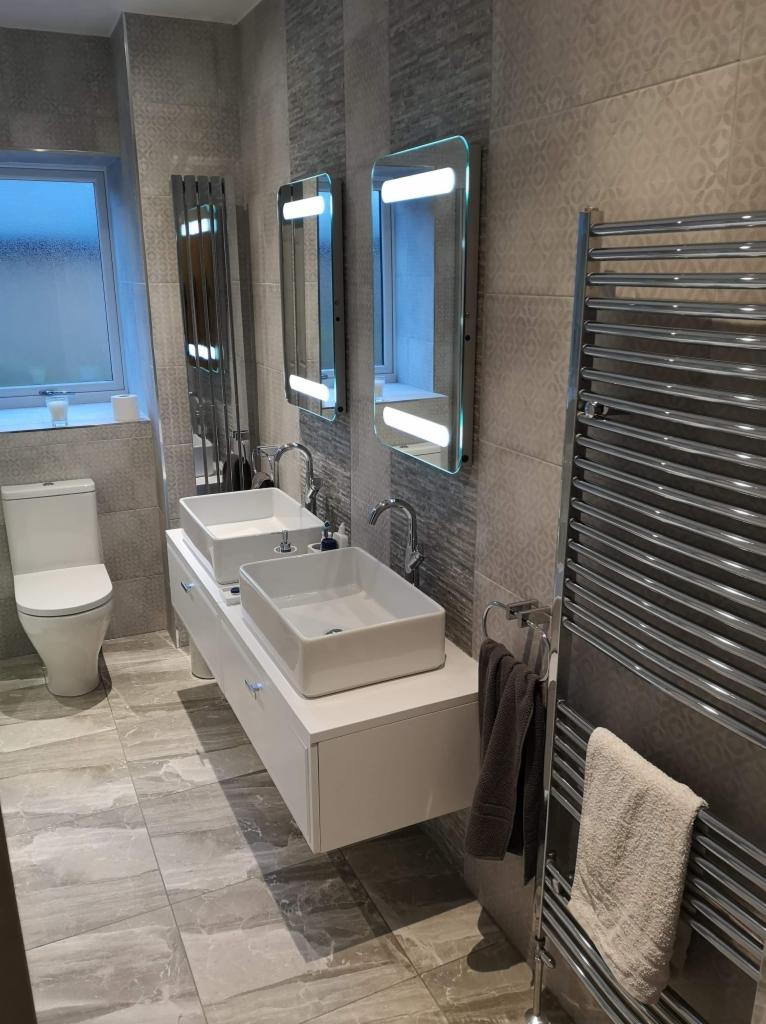 Wykończona łazienka w Jelcz-Laskowice z podwójną umywalką nablatową, grzejnikiem drabinkowym i podświetlanymi lustrami, płytki w odcieniach szarości na podłodze i ścianach.