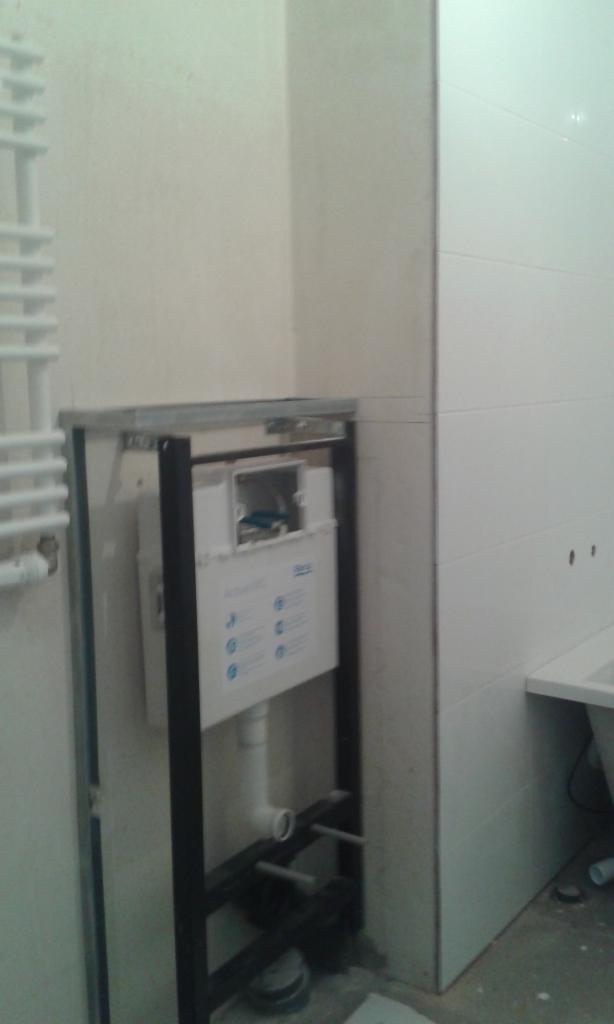 Stelaż podtynkowy WC zamontowany na ścianie w łazience z białymi płytkami, widoczne rury i instalacja wodno-kanalizacyjna.