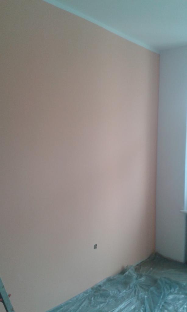 Ściana pomalowana na brzoskwiniowy kolor z widocznym gniazdkiem elektrycznym, podłoga zabezpieczona folią malarską, widoczny fragment drabiny.