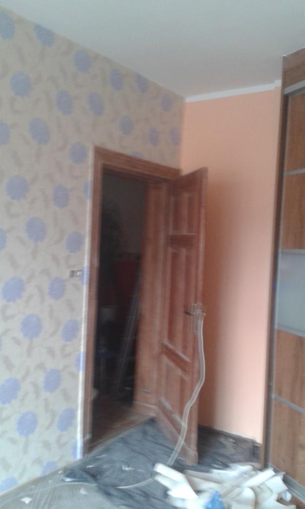 Ściana z tapetą w kwiaty, drewniana ościeżnica i częściowo otwarte drewniane drzwi, widoczne zabezpieczenia podłogi folią malarską i resztki styropianu po remoncie.