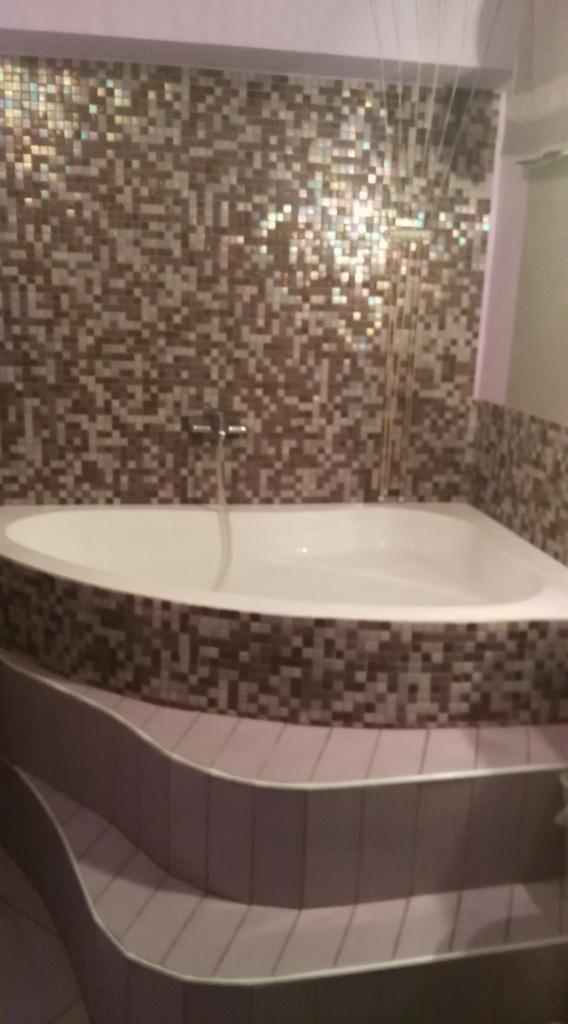 Biała wanna narożna z armaturą, obudowana mozaiką w odcieniach brązu i beżu, z dodatkowymi stopniami również wykończonymi mozaiką; ściana za wanną wyłożona identyczną mozaiką.