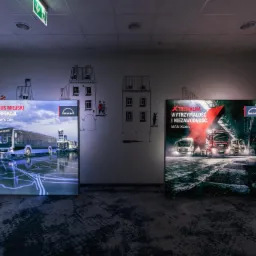 Dwa podświetlane panele reklamowe marki MAN prezentujące autobus miejski i ciężarówki, ustawione w pomieszczeniu na tle ściany z rysunkami budynków.