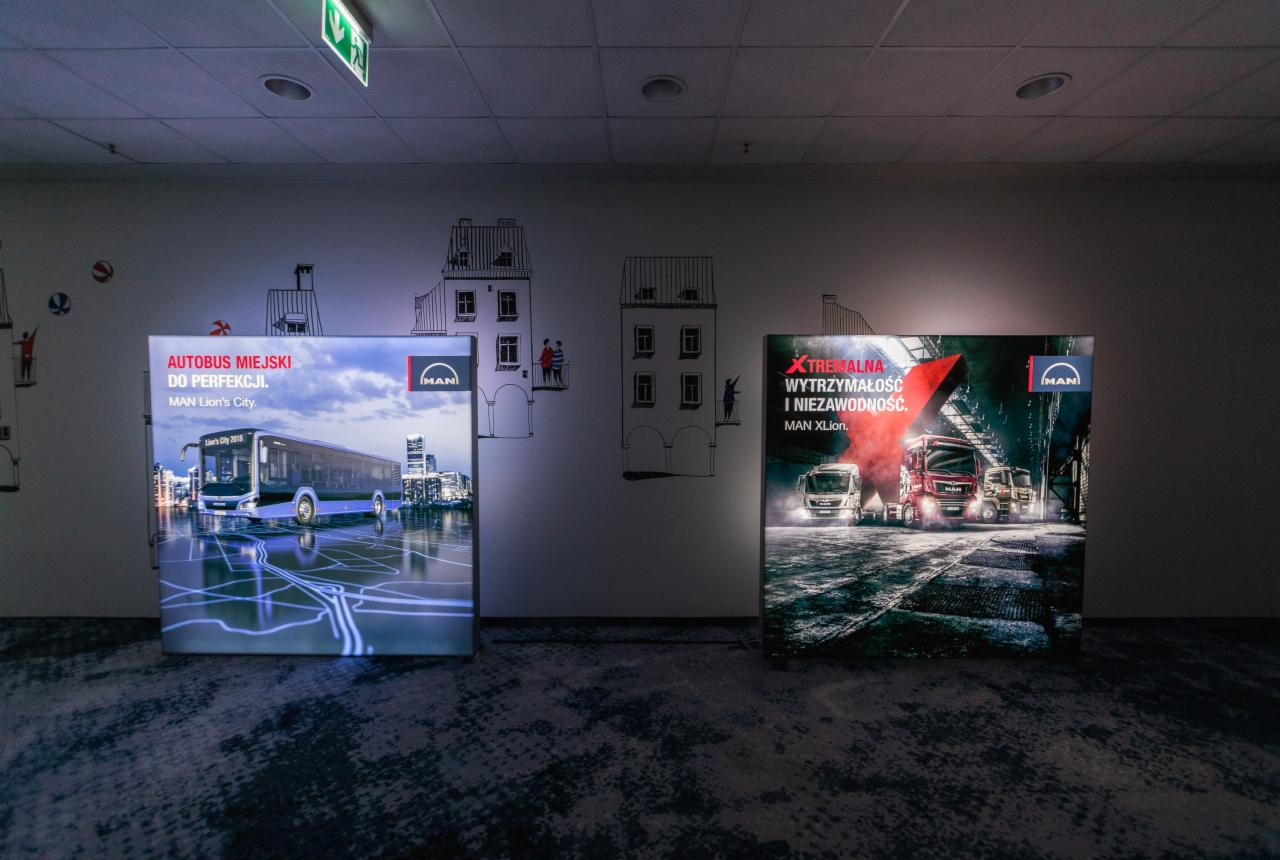 Dwa podświetlane panele reklamowe marki MAN prezentujące autobus miejski i ciężarówki, ustawione w pomieszczeniu na tle ściany z rysunkami budynków.