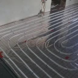 Białe rurki ogrzewania podłogowego ułożone na folii z czerwoną siatką, widoczne czerwone złączki, szare ściany w surowym stanie.