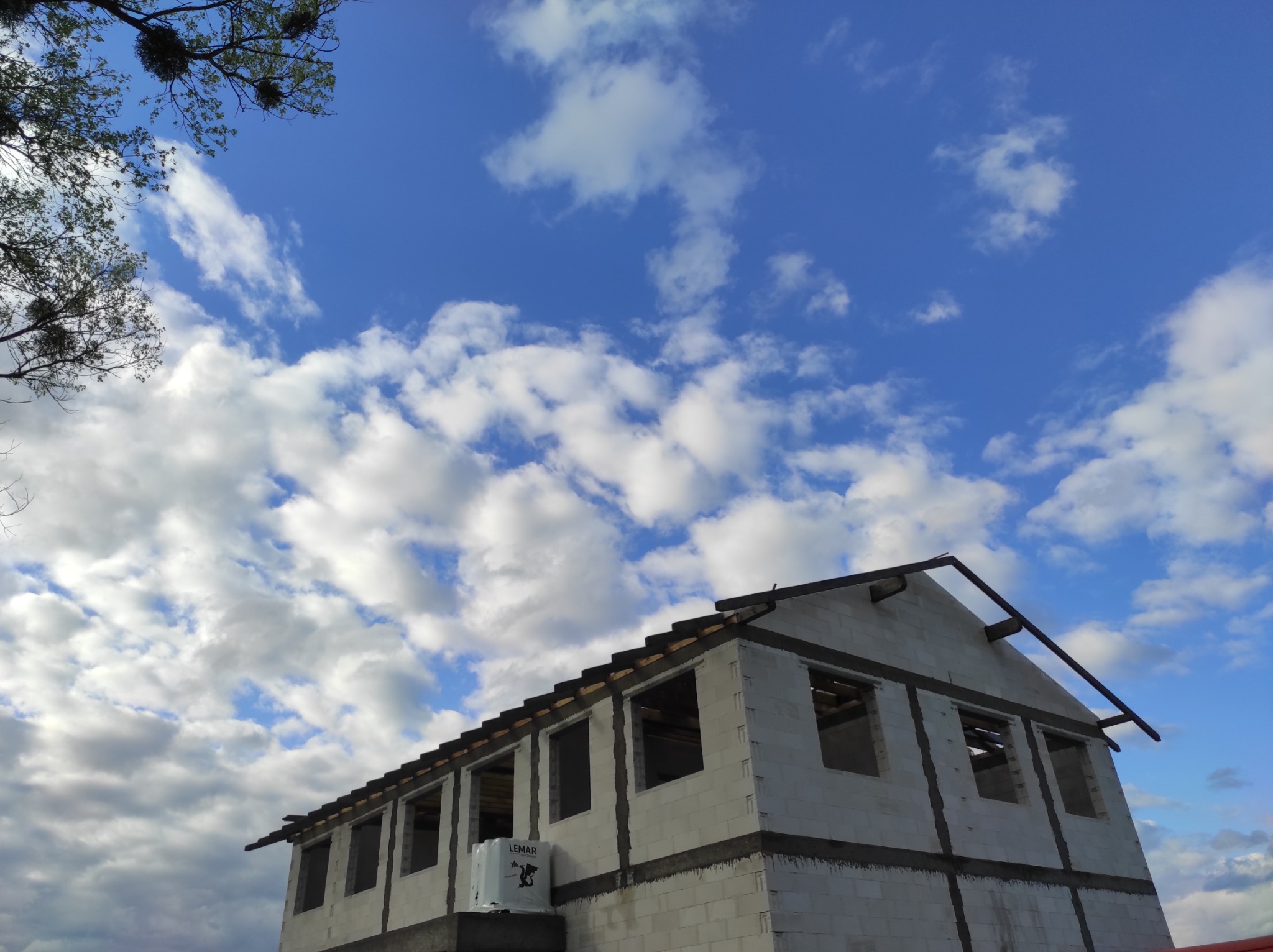 Surowy stan domu z białych bloczków, widoczne okna bez szyb, konstrukcja dachu w trakcie budowy, na tle błękitnego nieba z białymi chmurami.