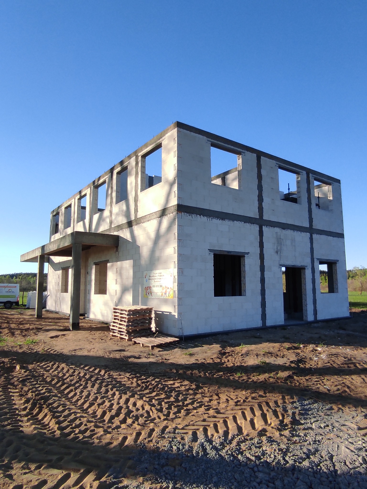 Dwukondygnacyjny dom w stanie surowym otwartym, zbudowany z bloczków betonowych, widoczny na tle błękitnego nieba i ziemi z wyraźnymi śladami opon ciężkiego sprzętu. Na ścianie budynku widoczna...