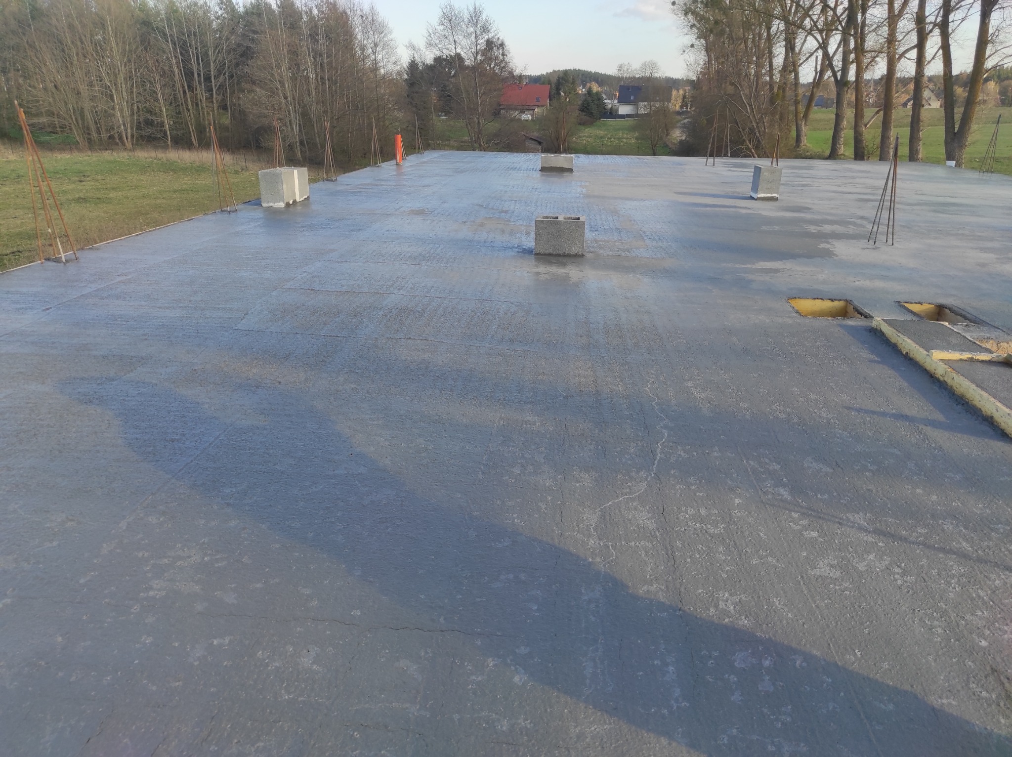 Zalana betonem płyta fundamentowa z wystającymi prętami zbrojeniowymi i bloczkami betonowymi, widok z góry na tle łąki i drzew