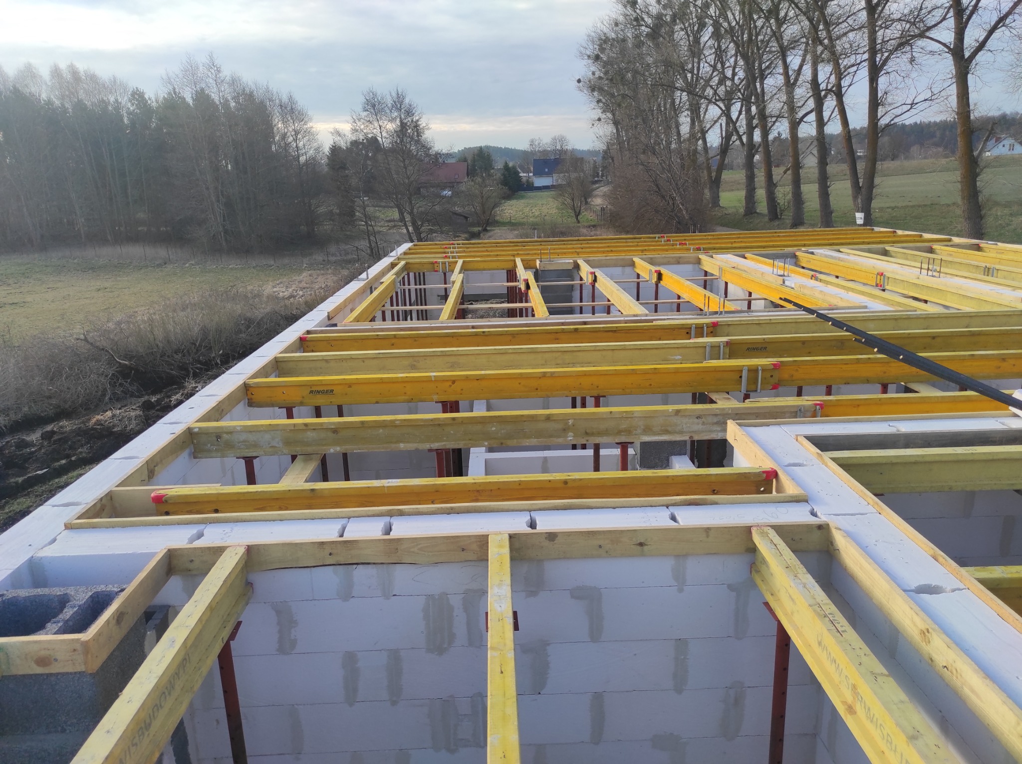 Wznoszenie ścian parteru domu z bloczków, widoczne żółte podpory szalunkowe, w tle łąka i drzewa.