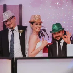 DJ z parą młodą pozują do zdjęcia w błyszczących kapeluszach podczas wesela, w tle dekoracje świetlne i rośliny.