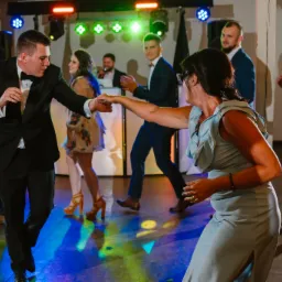 Roztańczona grupa gości weselnych na parkiecie pod oświetleniem scenicznym, z DJ-em w tle.