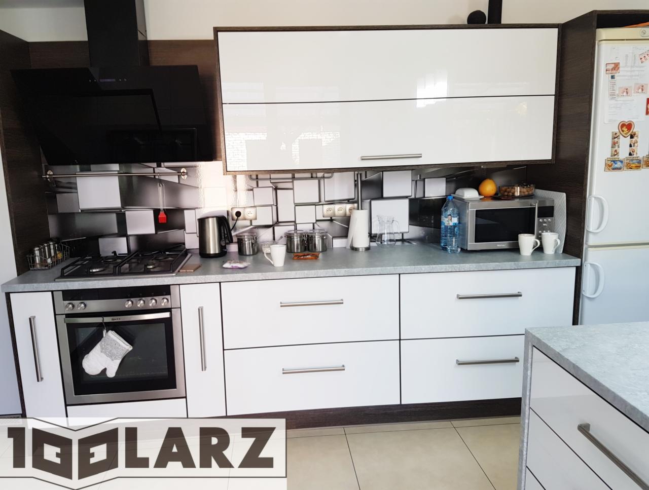 Nowoczesna kuchnia z białymi frontami szafek i szarym blatem, czarny okap i mozaika na ścianie, widoczne logo firmy w dolnej części kadru.