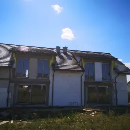 Nowy dach z ciemnej dachówki ceramicznej na dwukondygnacyjnym domu w stanie surowym otwartym, widoczne kominy, okna zabezpieczone folią, błękitne niebo z chmurami w tle.