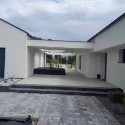 Usługi Remontowo-Wykończeniowe Dominik Ratajczak - Nowoczesny dom z białymi ścianami i szarą kostką brukową na podjeździe. Widoczny fragment ogrodu i zadaszone przejście. Minimalistyczny design.