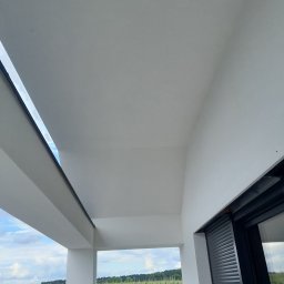 Usługi Remontowo-Wykończeniowe Dominik Ratajczak - Biała elewacja domu z widokiem na pole kukurydzy przez okno z roletami. Świeżo pomalowane ściany i sufit w jasnym odcieniu bieli. Minimalistyczny design.