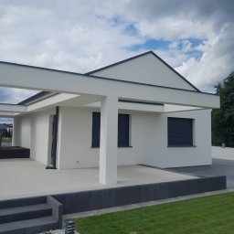 Usługi Remontowo-Wykończeniowe Dominik Ratajczak - Nowoczesny dom jednorodzinny z płaskim dachem i białymi ścianami. Widoczne szare rolety w oknach, zadbany trawnik i elementy małej architektury.