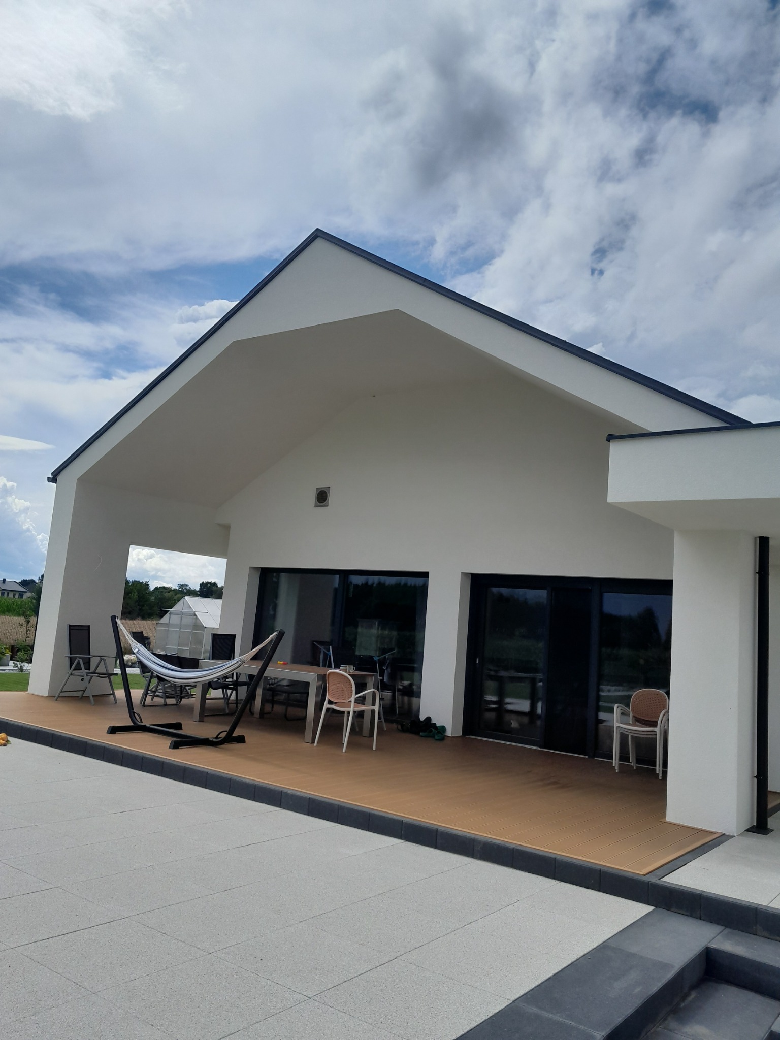 Nowoczesny dom z białymi ścianami i ciemnym dachem, taras z drewnianą podłogą, hamak i meble ogrodowe. Widok na ogród i błękitne niebo z chmurami.