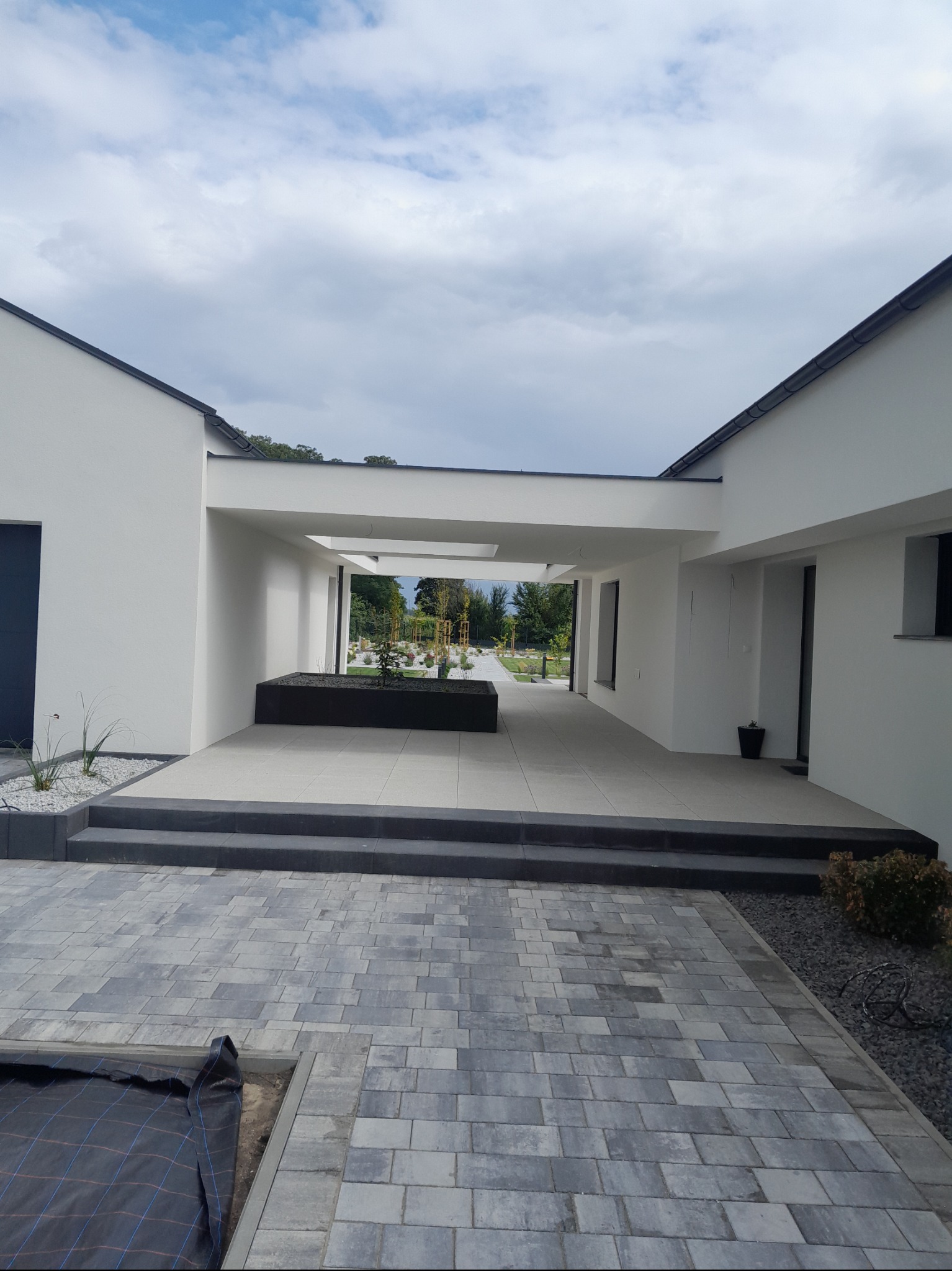 Nowoczesny dom z białymi ścianami i szarą kostką brukową na podjeździe. Widoczny fragment ogrodu i zadaszone przejście. Minimalistyczny design.