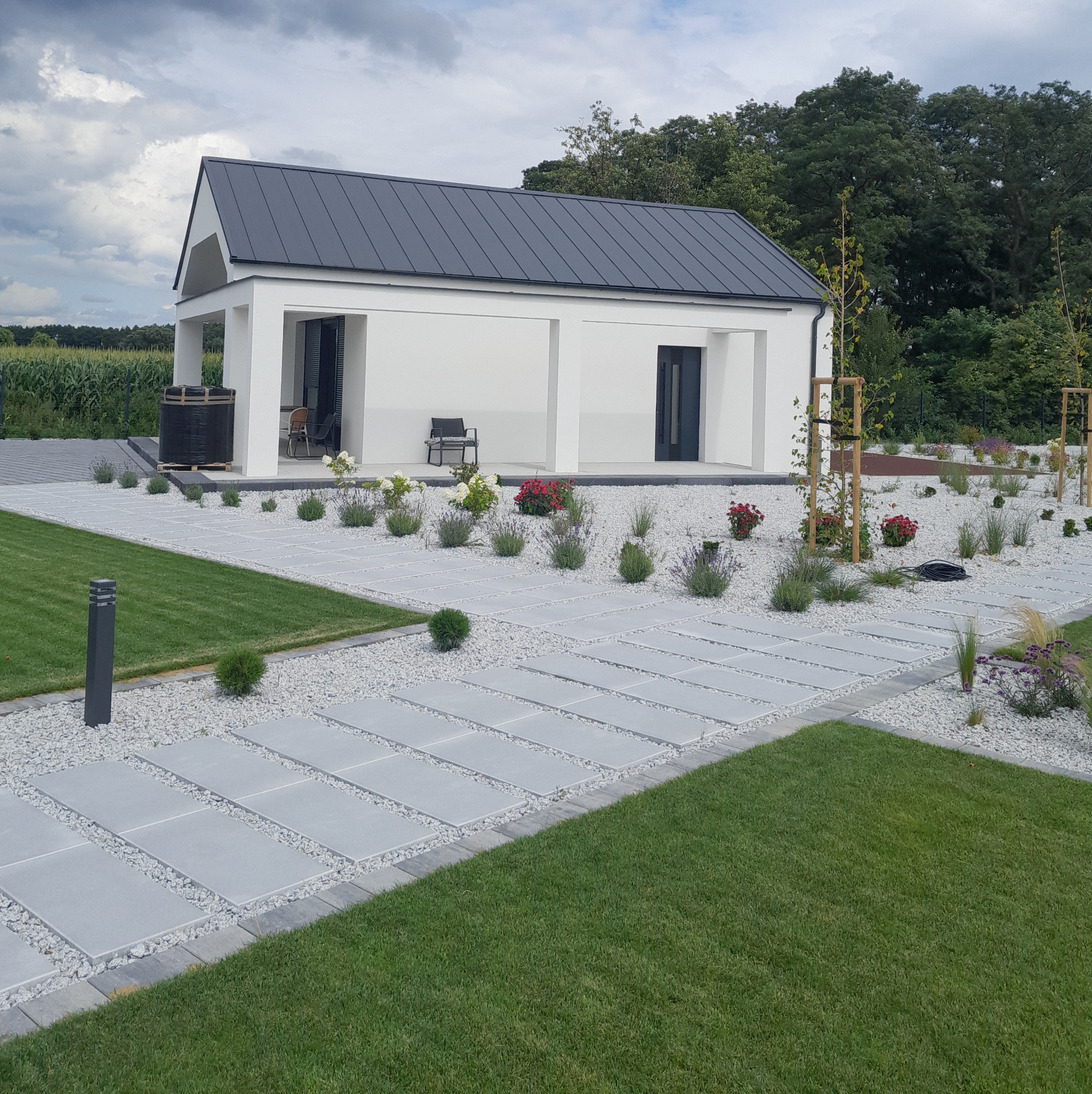 Nowoczesny dom jednorodzinny z białymi ścianami i grafitowym dachem, otoczony trawnikiem i ścieżką z płyt chodnikowych wysypaną białym kamieniem.