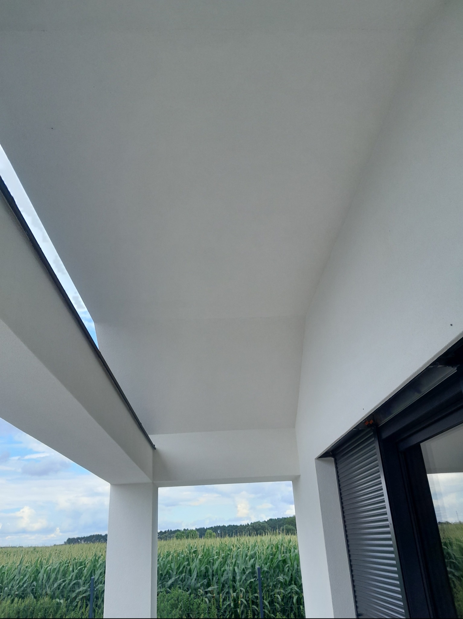 Biała elewacja domu z widokiem na pole kukurydzy przez okno z roletami. Świeżo pomalowane ściany i sufit w jasnym odcieniu bieli. Minimalistyczny design.