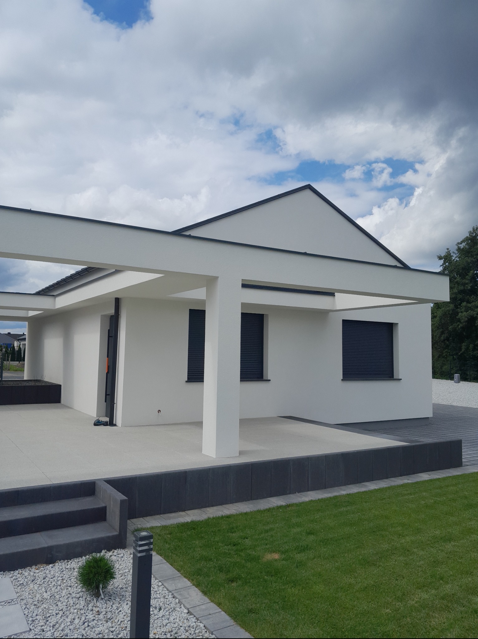Nowoczesny dom jednorodzinny z płaskim dachem i białymi ścianami. Widoczne szare rolety w oknach, zadbany trawnik i elementy małej architektury.