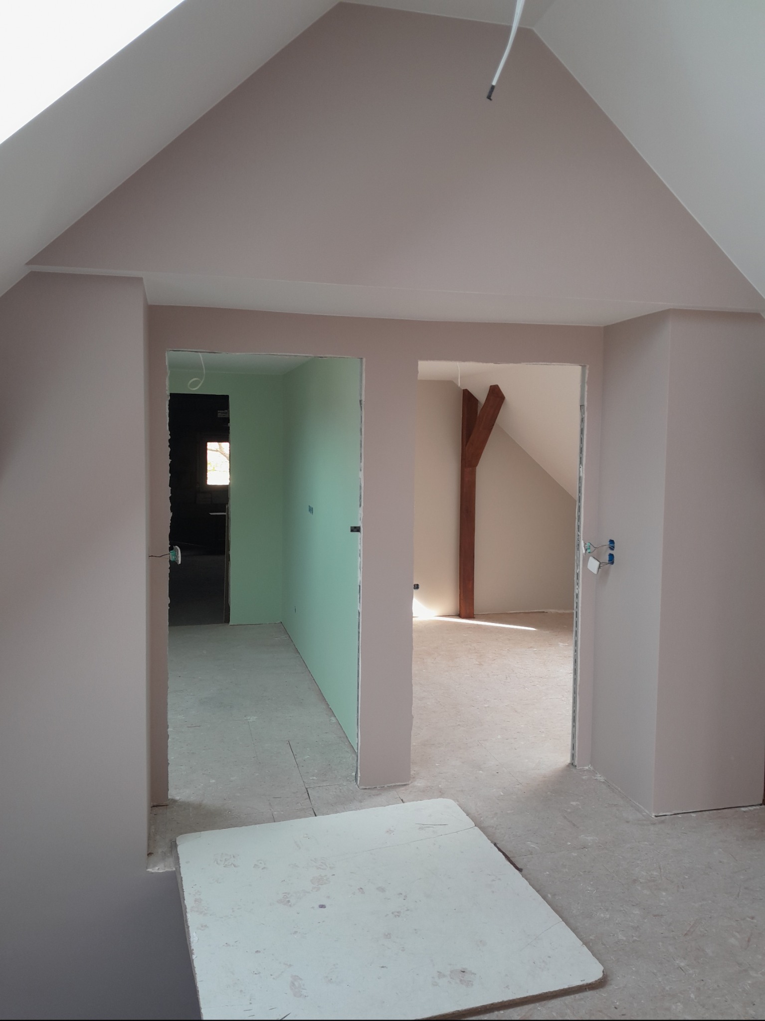 Poddasze w trakcie remontu: ściany pomalowane na beżowo, widoczne płyty karton-gips, drewniany element konstrukcyjny. Dwa otwarte przejścia do innych pomieszczeń.