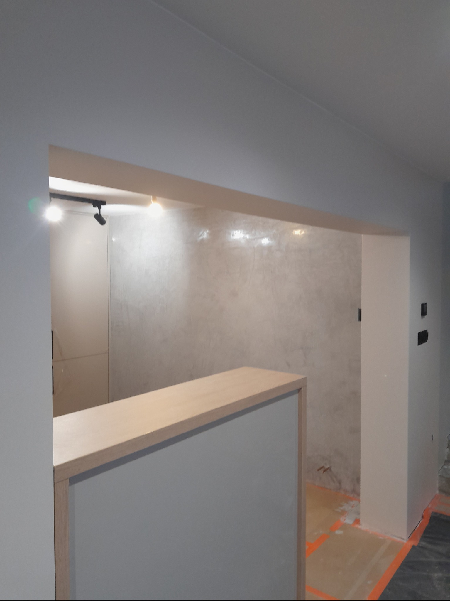 Wykończone wnętrze z dekoracyjną ścianą w odcieniach szarości, oświetleniem punktowym i elementami zabudowy. Widoczne zabezpieczenia podłogi wskazują na trwające prace.