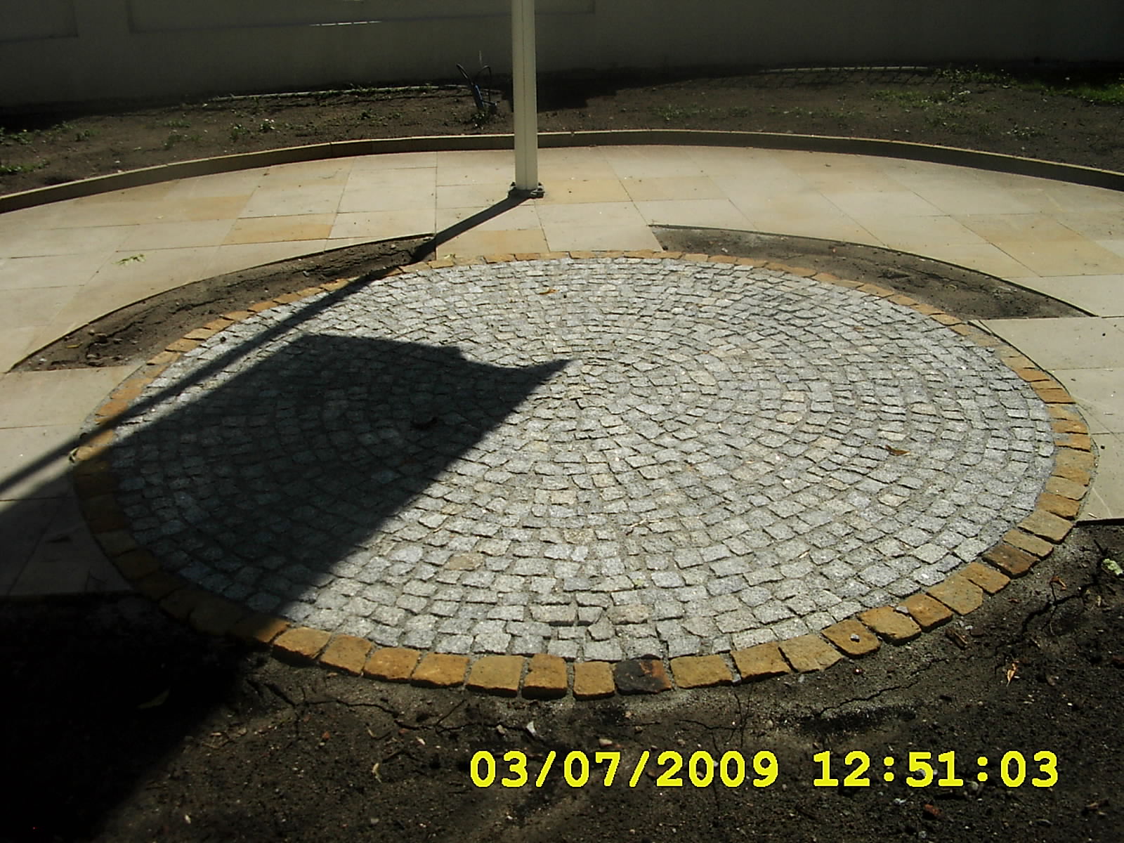 Okrągła nawierzchnia z kostki granitowej ułożona w spiralny wzór, obrzeże z cegieł, widoczny cień słupa, otoczona ziemią i płytami chodnikowymi.