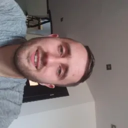 Uśmiechnięty mężczyzna w szarej koszulce, widoczny od klatki piersiowej w górę, z ciemnymi włosami i brodą, na tle jasnego pokoju z czarnymi elementami wyposażenia.