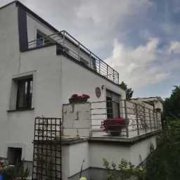 Dwupoziomowy dom z balkonami zabezpieczonymi poziomymi balustradami nierdzewnymi, z widocznymi donicami z czerwonymi kwiatami i dekoracyjnym drewnianym kołem na elewacji.